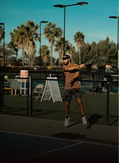 Pemain padel melompat memukul forehand di lapangan outdoor berlatar pohon palem